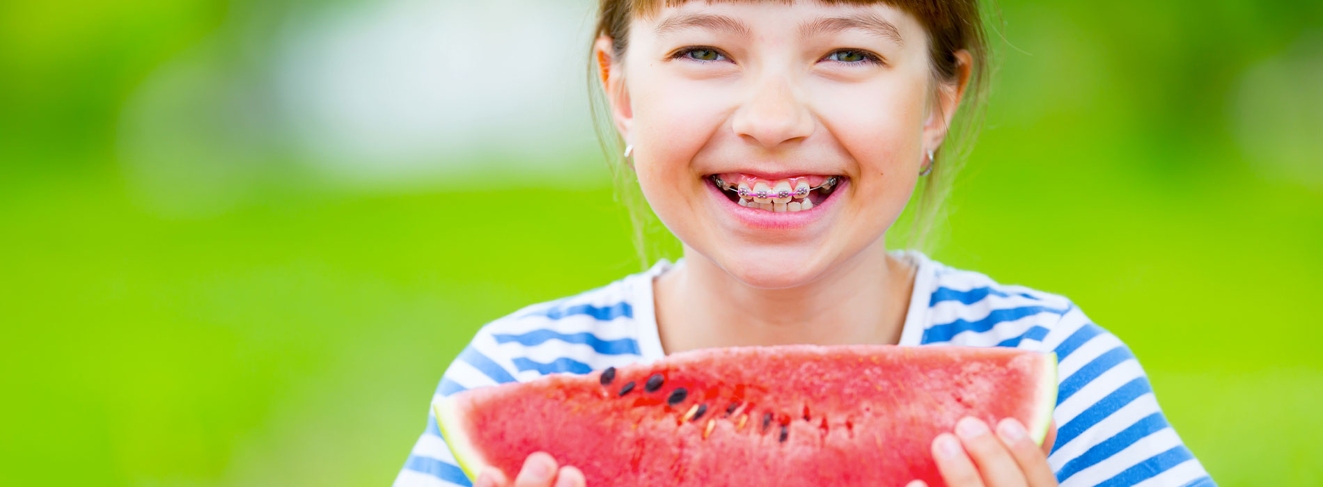 This is a photograph featuring a young girl holding a watermelon with a joyful expression, set against an outdoor background with blurred greenery.