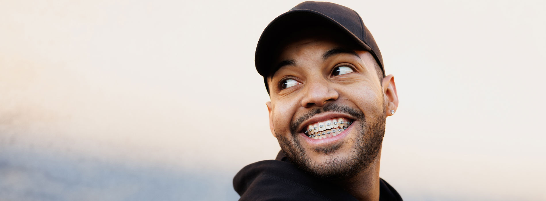 The image shows a man with a beard and mustache smiling at the camera, wearing a black baseball cap, with a clear sky background.