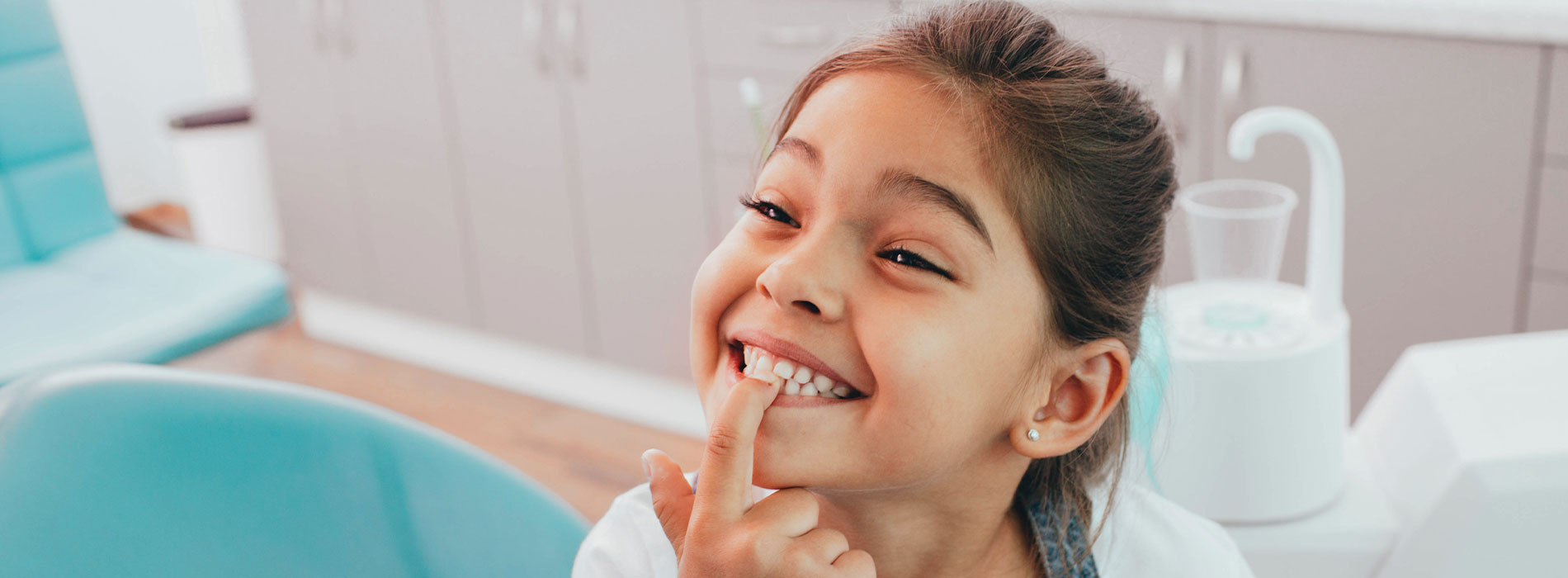 A young girl with a big smile, sitting in a dental chair, looking directly at the camera.