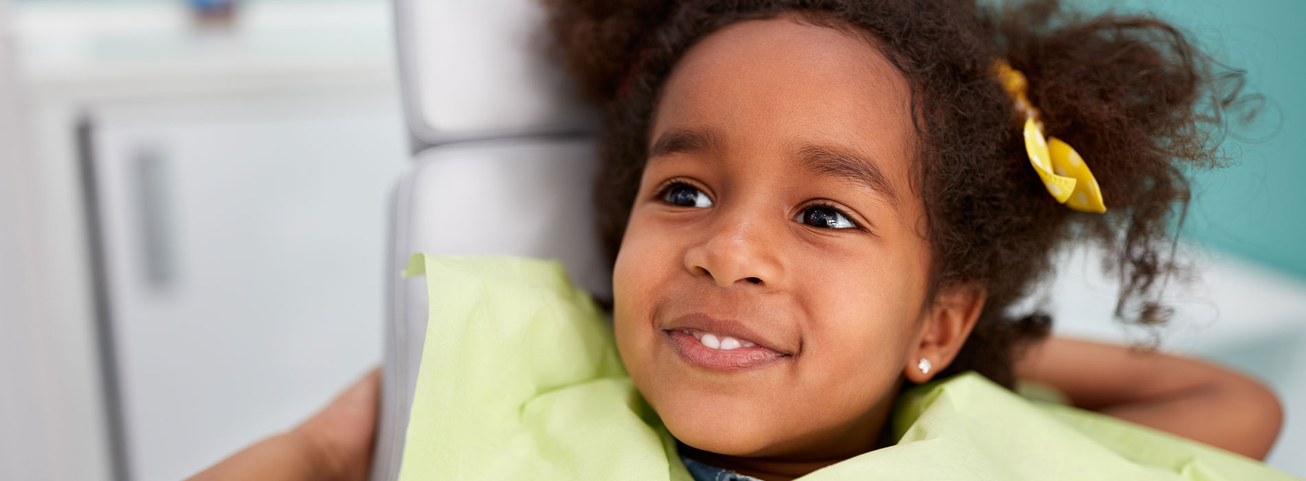 A young girl with dark hair smiling at the camera while sitting on a dental chair.