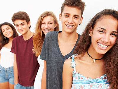 The image shows a group of five young people posing together, with two males on the left and three females on the right, all smiling and looking towards the camera. They are outdoors during the daytime under clear skies.